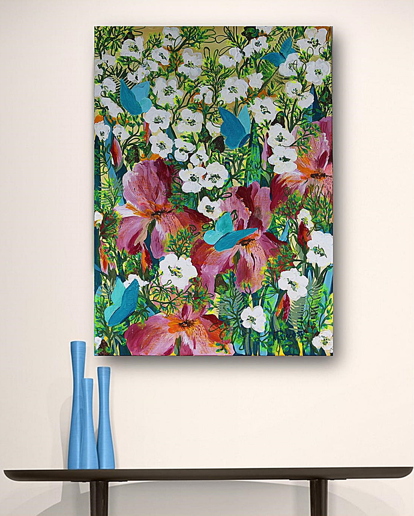 A vertical original canvas wall art  featuring a colorful floral painting with white flowers, pink and purple blossoms, green foliage, and blue butterflies, displayed on a beige wall above a dark console table with blue vases.
