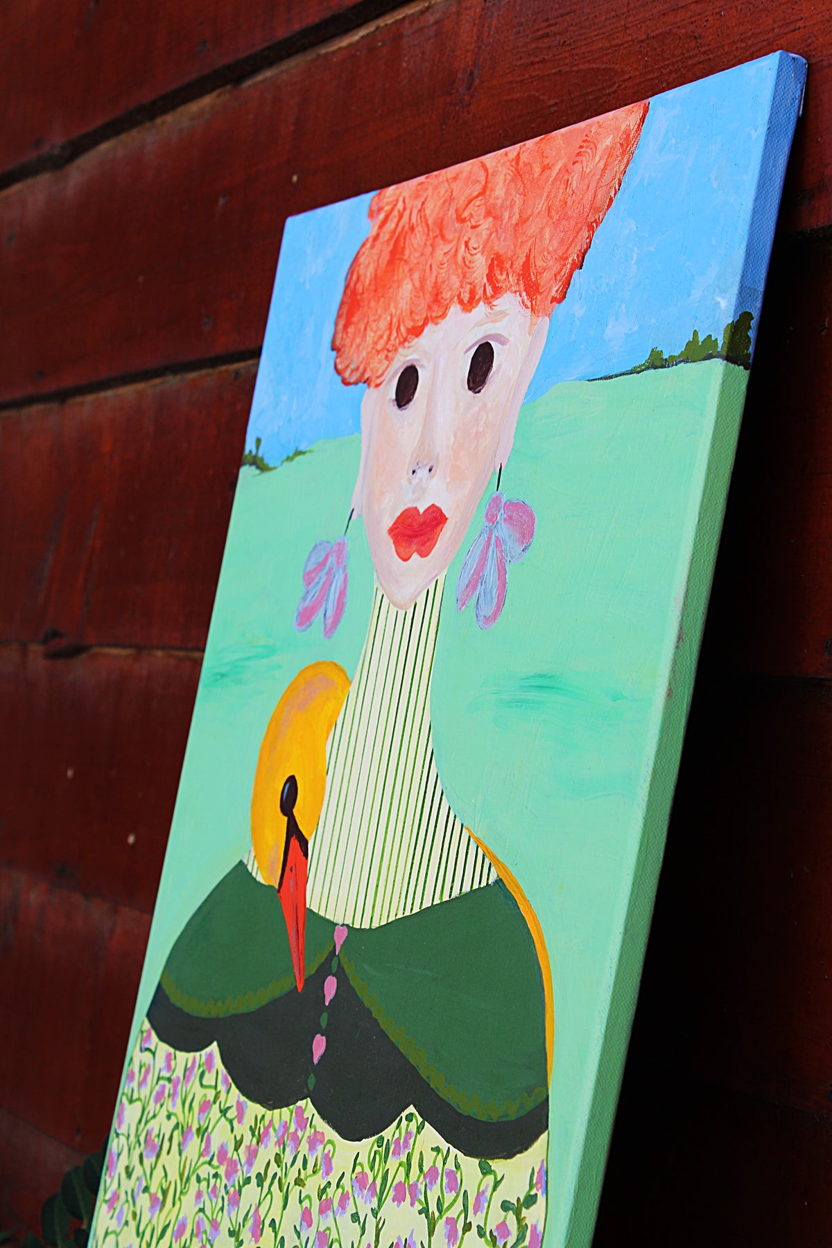 Lateral view of a colorful painted canvas leaning against a dark red wooden wall. The artwork depicts a stylized figure with bright orange hair, a striped cream neck, green clothing, and a light blue sky and grassy background.